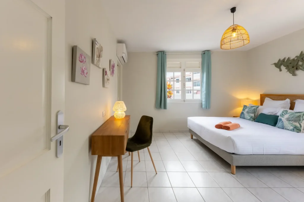 Chambre lumineuse avec lit double, bureau, chaise et fenêtre aux rideaux turquoise.