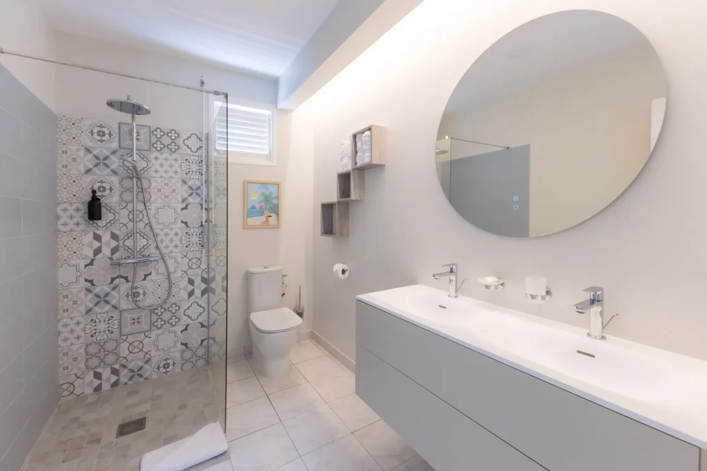 Salle de bain moderne avec douche, lavabo double et miroir rond. Décor épuré et lumineux.
