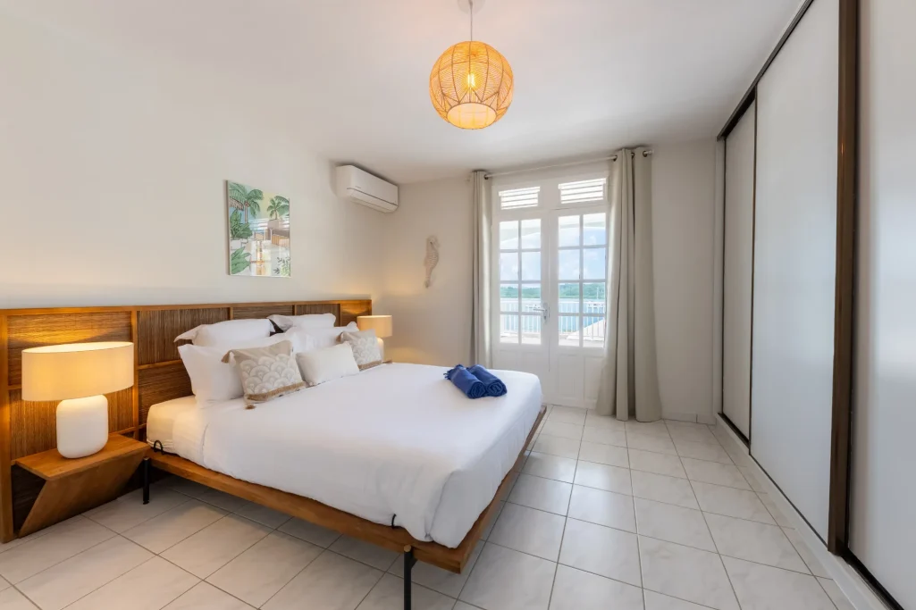 Chambre lumineuse avec lit double, tapis, et grandes fenêtres. Décoration moderne et épurée.