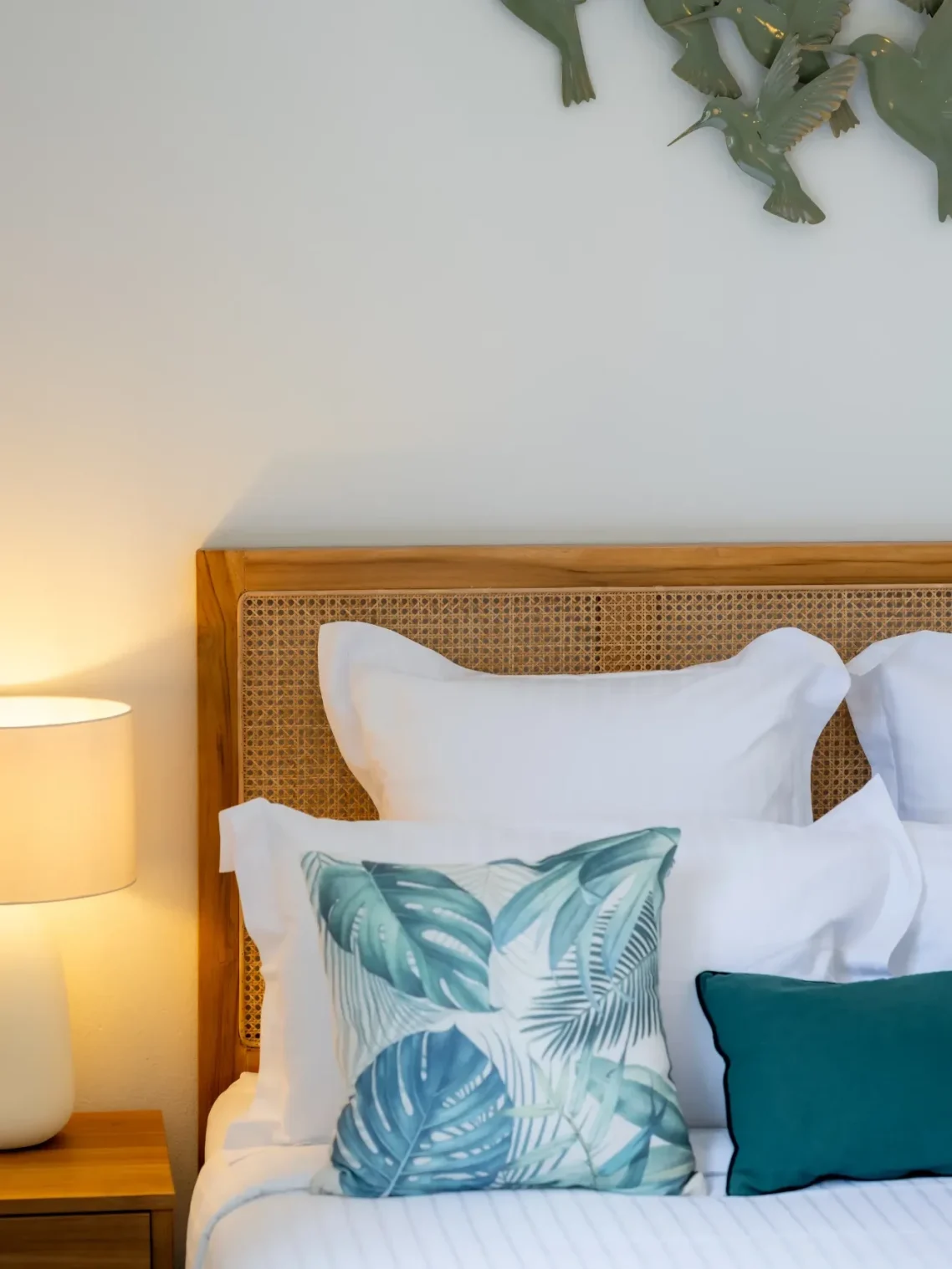 Lit de chambre avec oreillers blancs et coussins aux motifs tropicaux, lampe allumée.